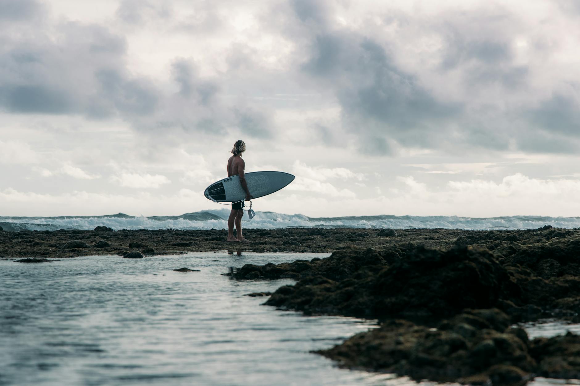 Surfer z deską na skalistej plaży w Indonezji przy pochmurnym niebie